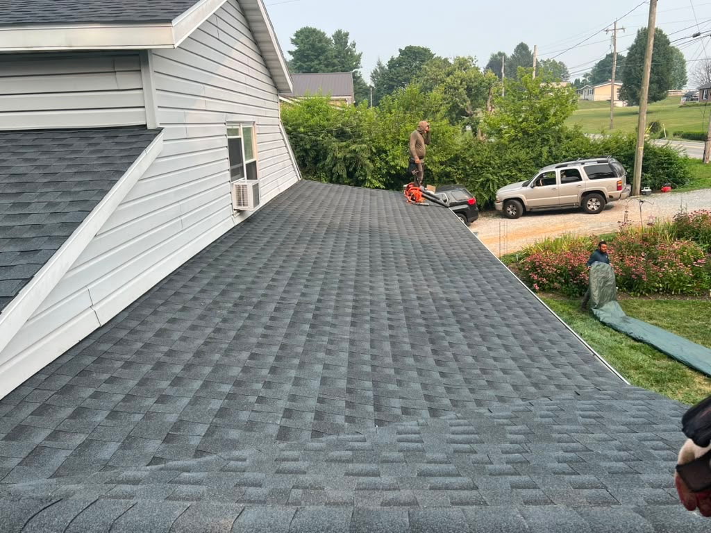 New charcoal architectural shingle roof on a white two-story home with a crew member clearing debris near the ridge