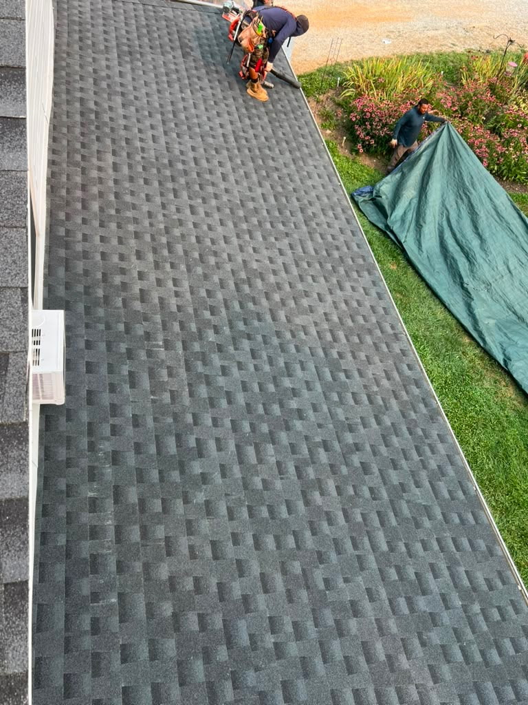 Long run of new charcoal shingles down a steep roof with a crew member at the top and a debris tarp spread on the lawn