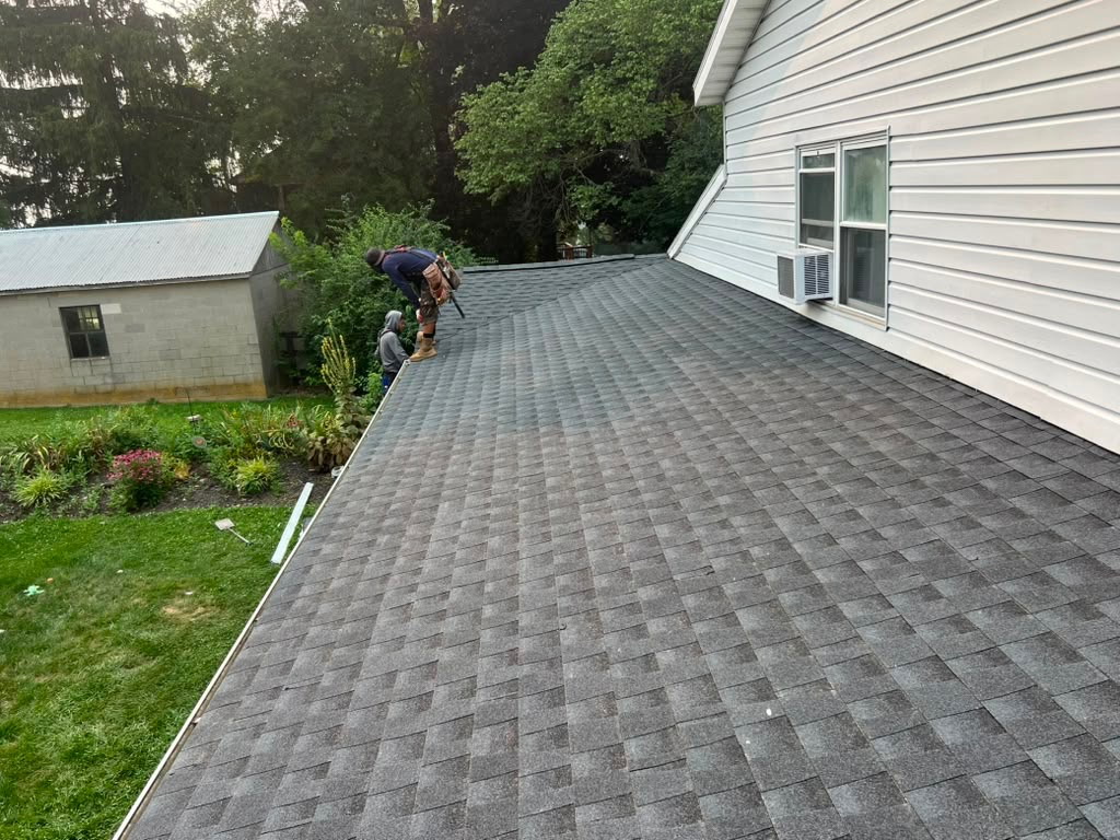 Dallastown crew member finishing the edge of a new charcoal architectural shingle roof on a white-sided home above a flower garden