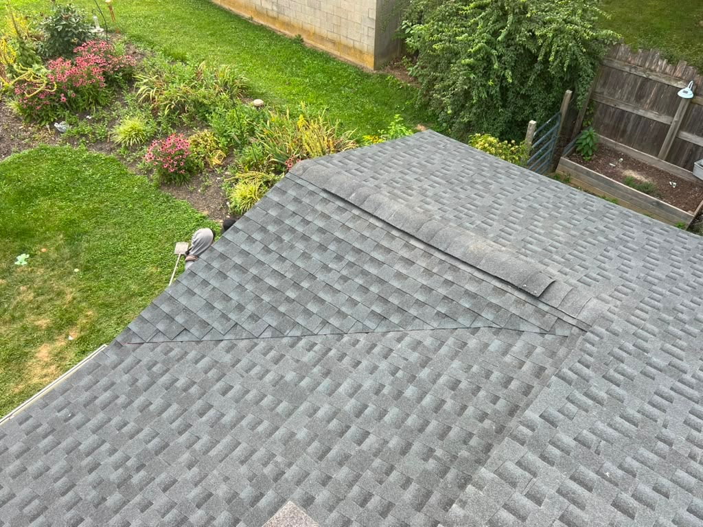 Grey architectural shingle hip roof with the ridge cap being installed, overlooking a backyard flower garden
