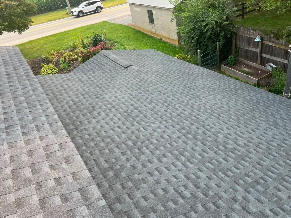 Aerial view of a new grey architectural shingle roof with a country road, outbuilding, and garden beds beyond